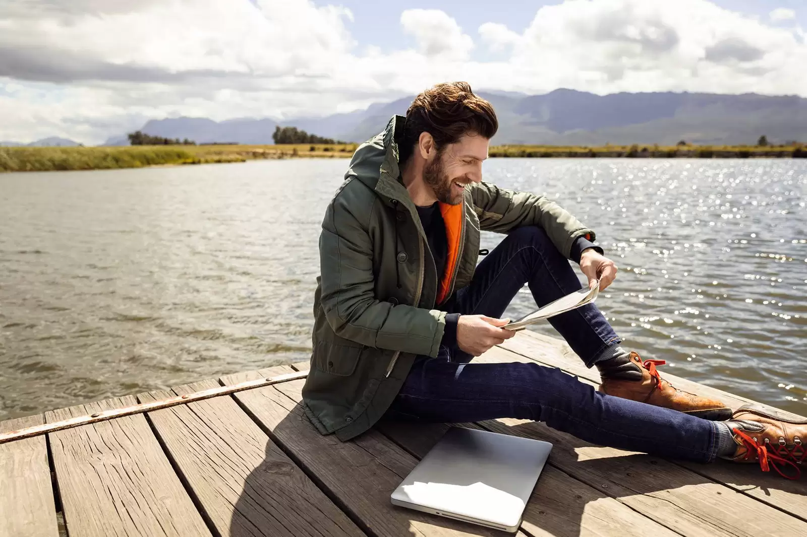 Hombre joven con un ordenador leyendo unos papeles sentado en un muelle con un lago de fondo