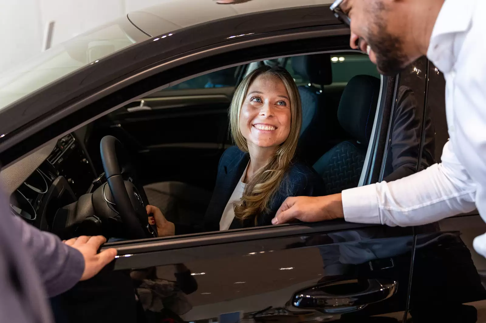 Mujer sonriendo sentada al volante de un Volkswagen mirando por la ventanilla a dos hombres