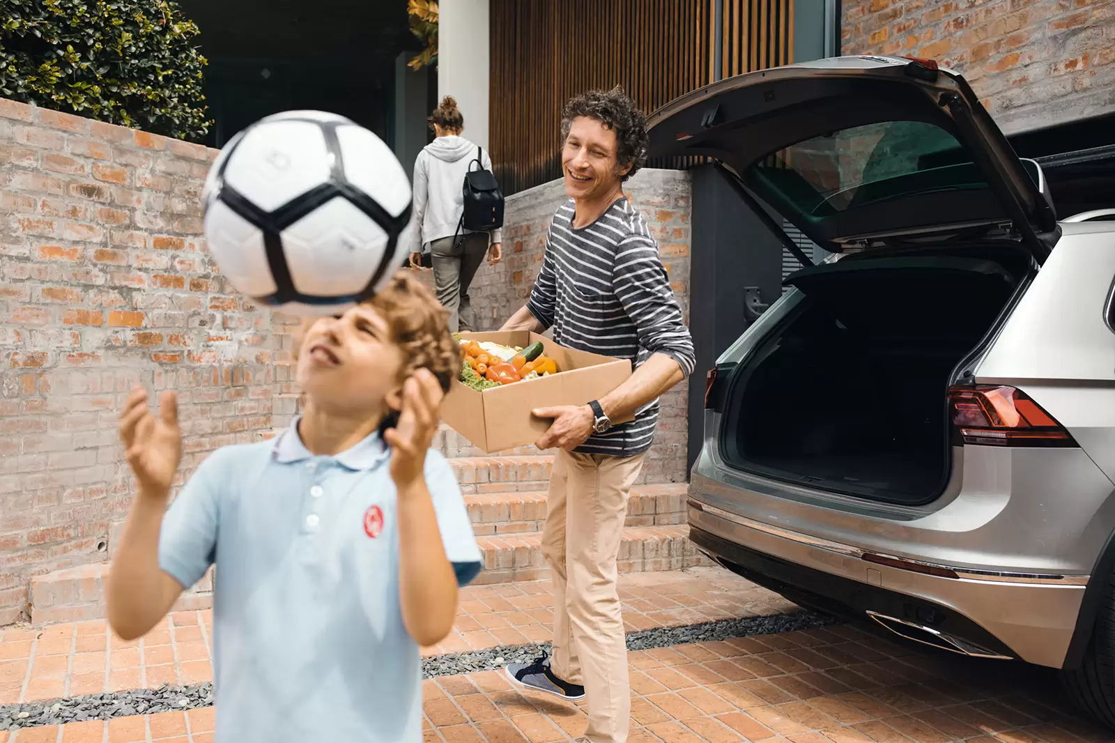 Niño jungando con una pelota mientras lo mira su padre que baja una caja de un coche