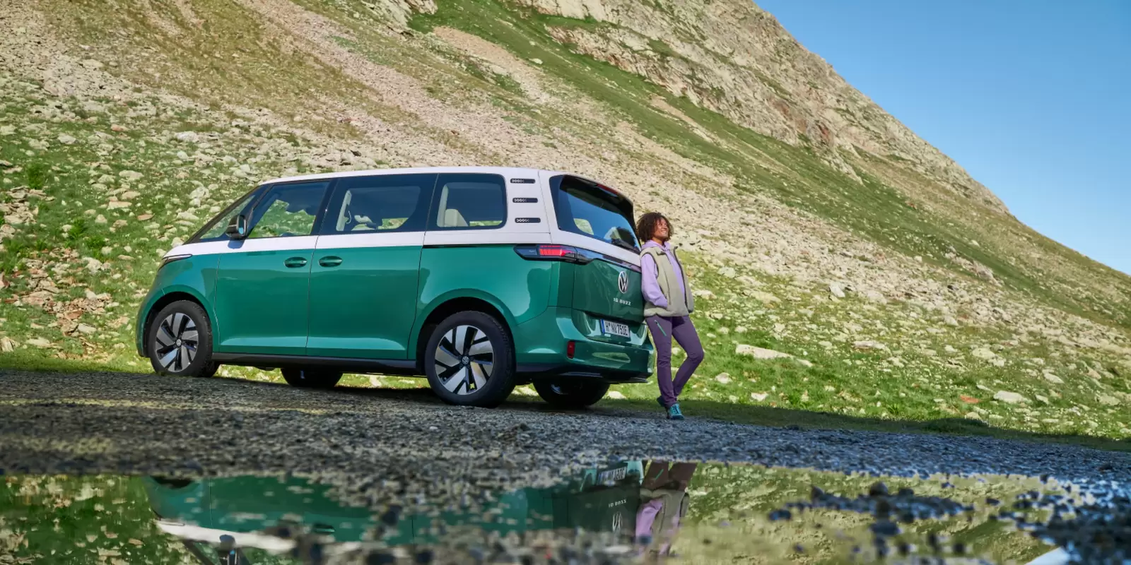 Una chica apoyada en  un Volkswagen ID. Buzz de Renting bicolor verde y blanco en la montaña